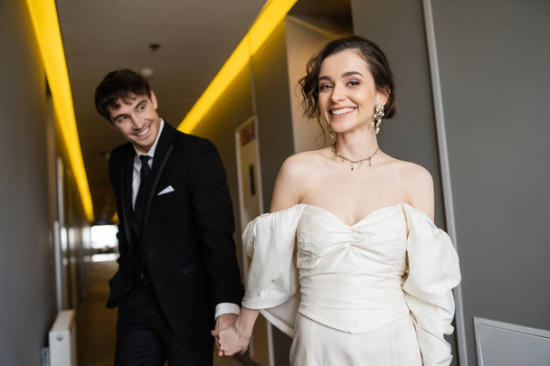blurred and cheerful man in black suit holding hands with gorgeous bride in white wedding dress while smiling and walking together in corridor of modern hotel, honeymoon   - Photo, Image