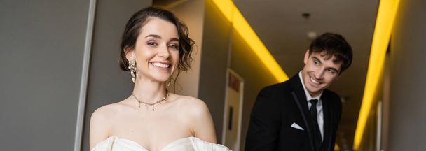 blurred and cheerful man in black suit looking at gorgeous bride in white wedding dress while smiling and walking together in corridor of modern hotel, banner  - Foto, imagen