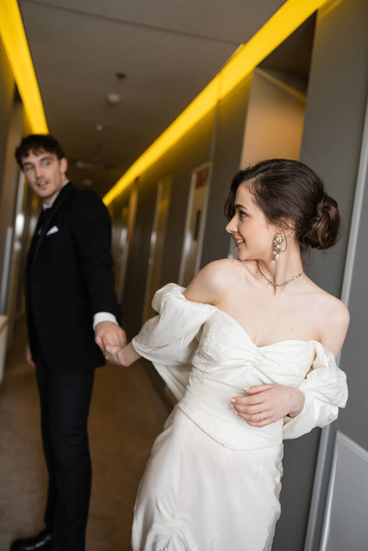 gorgeous bride in white wedding dress holding hands with blurred and cheerful groom in black suit while smiling and walking together in corridor of modern hotel  - Foto, imagen