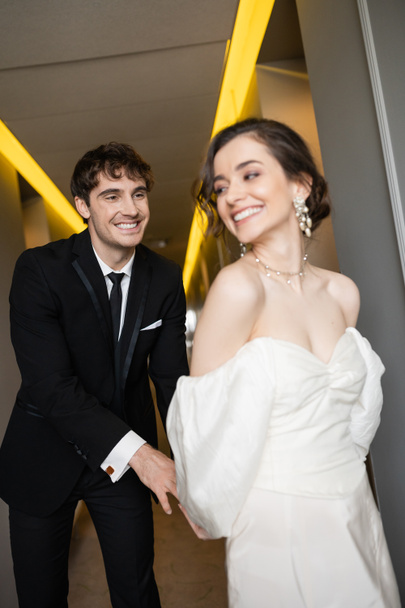 Χαρούμενος γαμπρός με μαύρο κοστούμι γαργαλάει απολαυστική νύφη με λευκό νυφικό ενώ χαμογελά και περπατά μαζί στο διάδρομο του σύγχρονου ξενοδοχείου, ευτυχισμένοι νεόνυμφοι στο μήνα του μέλιτος  - Φωτογραφία, εικόνα