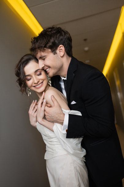cheerful groom in black suit embracing delightful bride in white wedding dress while smiling and standing together in hallway of modern hotel, happy newlyweds on honeymoon  - Foto, imagen