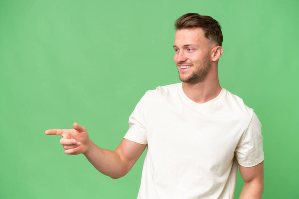 Jonge blonde blanke man over geïsoleerde achtergrond wijzend vinger naar de zijkant en presenteren een product - Foto, afbeelding