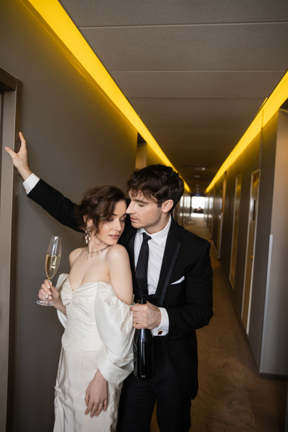 young groom in black suit leaning towards wall and holding bottle near stunning bride with glass of champagne while standing together in hallway of modern hotel, newlyweds on honeymoon  - Foto, afbeelding