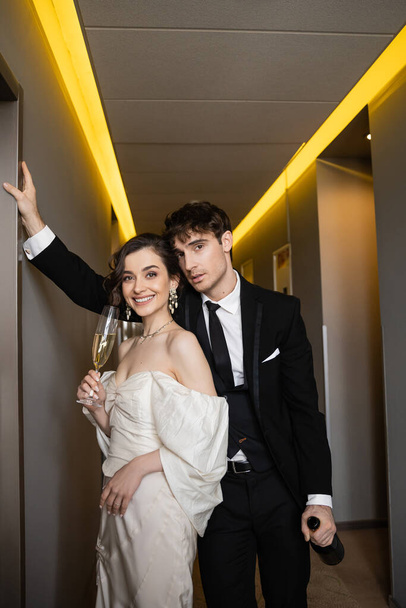 good looking groom in black formal wear leaning towards wall and holding bottle near happy bride with glass of champagne while standing together in hallway of modern hotel  - 写真・画像