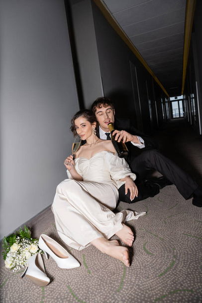 groom in black suit drinking from bottle and sitting near gorgeous bride with glass of champagne next to bridal bouquet and high heels on floor in hall of modern hotel  - 写真・画像