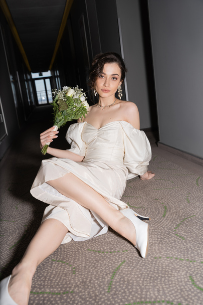mladá a brunetka nevěsta v bílých svatebních šatech při pohledu na kameru při držení svatební kytice s květinami a posezení na podlaze chodby v moderním hotelu  - Fotografie, Obrázek