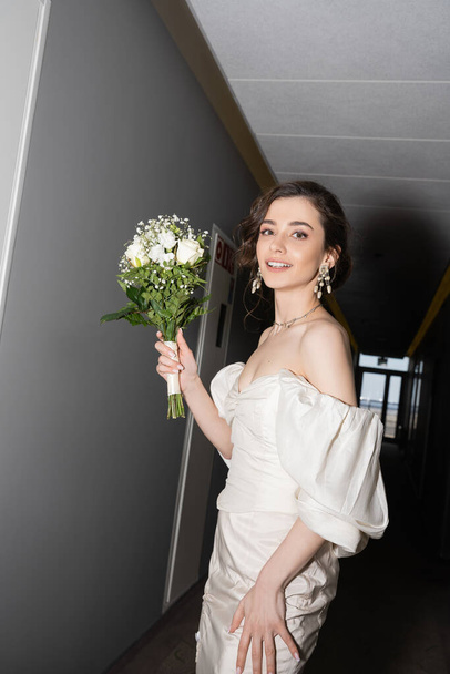 veselá a brunetka mladá nevěsta v bílých šatech s úsměvem při držení svatební kytice s květinami a při pohledu na kameru v hale v moderním hotelu, krásná nevěsta na svatební den - Fotografie, Obrázek