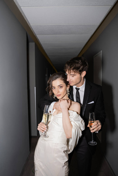 young and brunette bride in white wedding dress and handsome groom holding glasses of champagne while standing together in hallway of hotel, newlyweds on honeymoon  - Foto, immagini