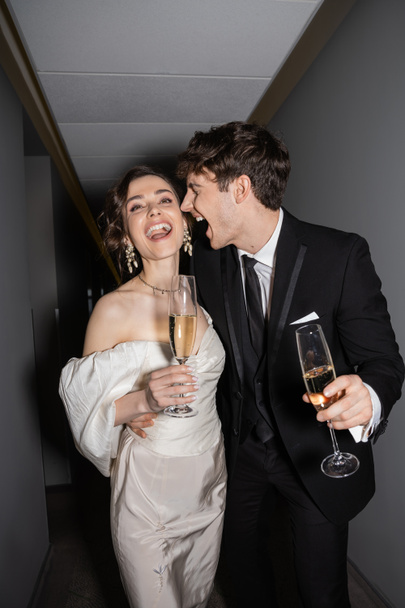 excited groom hugging young and brunette bride in white wedding dress and holding glasses of champagne while standing and smiling together in hallway of hotel, newlyweds on honeymoon  - Фото, зображення