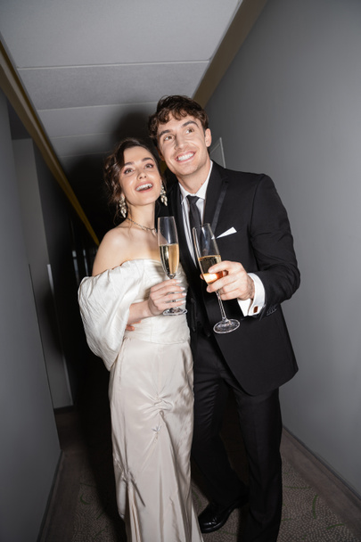 joyful groom hugging young and brunette bride in white wedding dress and holding glasses of champagne while standing and smiling together in hallway of hotel  - Фото, зображення