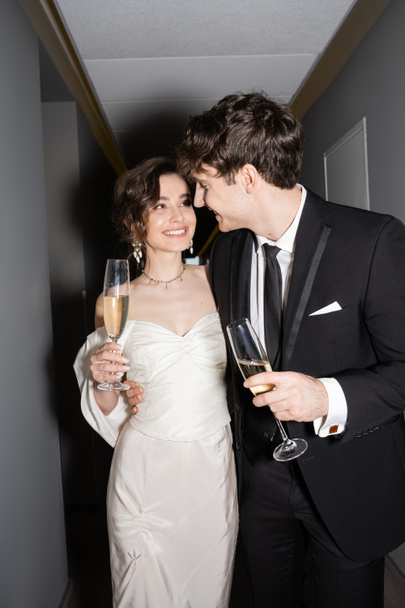 cheerful groom hugging young and happy bride in white wedding dress and holding glasses of champagne while standing and smiling together in corridor of hotel, newlyweds on honeymoon  - Foto, Bild
