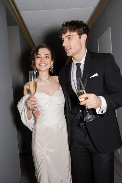 handsome groom hugging young and happy bride in white dress and holding glasses of champagne while standing and smiling together in corridor of hotel  - Foto, afbeelding