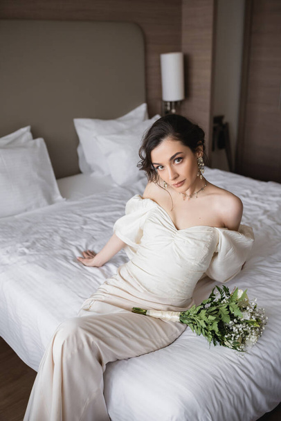mladá nevěsta v bílých šatech a luxusní šperky sedí na posteli vedle svatební kytice s květinami a při pohledu na kameru v moderní ložnici v hotelovém pokoji na svatební den  - Fotografie, Obrázek
