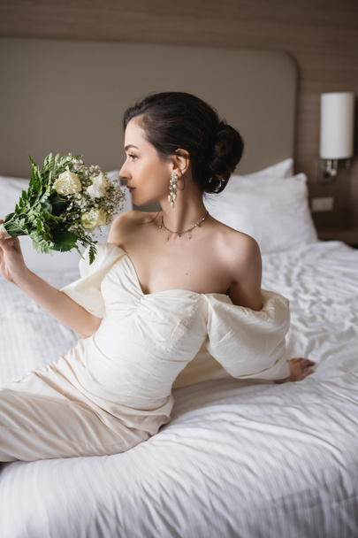 mladá nevěsta v bílých šatech a luxusní šperky sedí na posteli a voní svatební kytice s květinami, zatímco odvrací pohled v moderní ložnici v hotelovém pokoji na svatební den  - Fotografie, Obrázek
