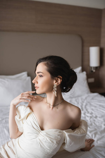 boční pohled na okouzlující mladou nevěstu v bílých šatech a luxusní šperky s perlovými náušnicemi a náhrdelníkem odvracející se v moderní ložnici v hotelovém pokoji v den svatby  - Fotografie, Obrázek