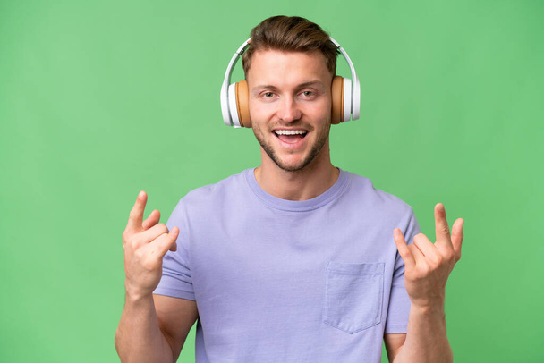 Молода блондинка білий чоловік над ізольованим фоном слухає музику, роблячи рок жест
 - Фото, зображення