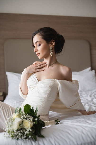 okouzlující mladá nevěsta v bílých šatech a luxusní šperky sedí na posteli vedle svatební kytice s květinami v moderní ložnici hotelového pokoje na svatební den  - Fotografie, Obrázek