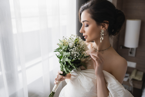 side view of elegant young bride in white dress and luxurious jewelry with pearl earrings holding bridal bouquet in modern hotel room on wedding day  - Photo, Image