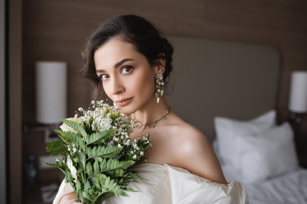 czarująca młoda panna młoda w białej sukience i luksusowej biżuterii trzymając bukiet ślubny z kwiatami i patrząc na aparat fotograficzny w nowoczesnej sypialni w pokoju hotelowym w dniu ślubu  - Zdjęcie, obraz