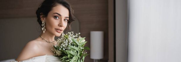 okouzlující mladá žena v bílých svatebních šatech a luxusní šperky držení svatební kytice s květinami a při pohledu na kameru v moderní ložnici v hotelovém pokoji, banner  - Fotografie, Obrázek