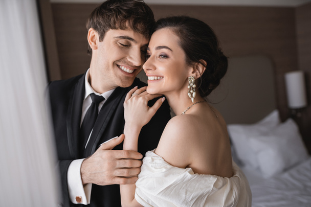 radosna młoda panna młoda w biżuterii i sukni ślubnej przytulając ramię wesołego pana młodego w klasycznym stroju formalnym stojąc razem w nowoczesnym pokoju hotelowym po ceremonii  - Zdjęcie, obraz