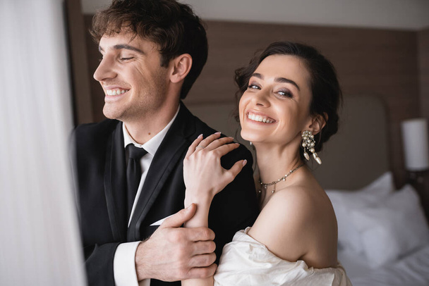 radostná nevěsta v elegantních špercích a svatebních šatech objímající rameno šťastného ženicha v klasickém formálním oblečení při společném stání v moderním hotelovém pokoji po obřadu  - Fotografie, Obrázek