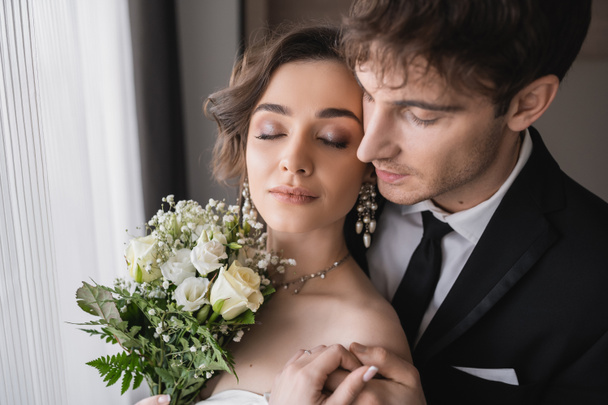 pretty bride in jewelry, white dress with bridal bouquet holding hands with groom in classic formal wear while standing together with closed eyes in modern hotel room after wedding ceremony  - Fotó, kép