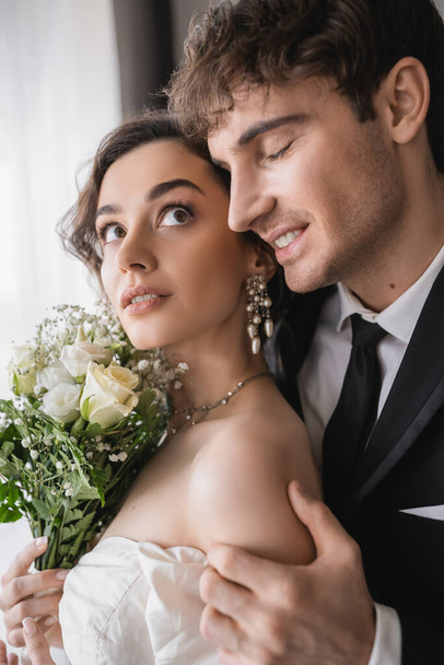 šťastný ženich v klasické formální oblečení dotýkat ruku krásné mladé nevěsty v šperky, bílé šaty s svatební kytice, zatímco stojí spolu v moderním hotelovém pokoji po svatebním obřadu  - Fotografie, Obrázek