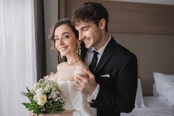 結婚式の後、現代のホテルの部屋に一緒に立っている間、古典的な正式な摩耗で幸せな新郎は、ジュエリーでエレガントな若い花嫁の肩に触れて、ブライダル花束と白いドレス  - 写真・画像