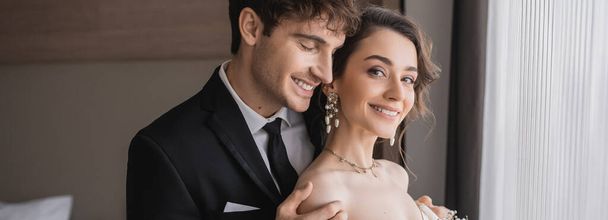 šťastný ženich v klasickém formálním oděvu dotýkající se ramene elegantní a veselé mladé nevěsty v špercích s perlami při společném postávání v moderním hotelovém pokoji po svatebním obřadu, banner  - Fotografie, Obrázek