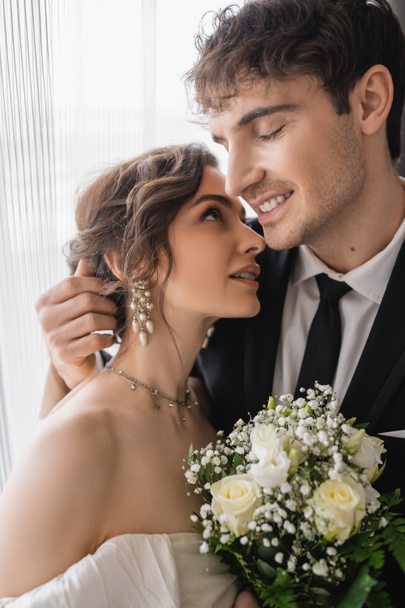 vrolijke bruidegom in klassieke formele dragen knuffelen gelukkige bruid in sieraden, witte jurk met bruids boeket terwijl samen staan in moderne hotelkamer na huwelijksceremonie  - Foto, afbeelding