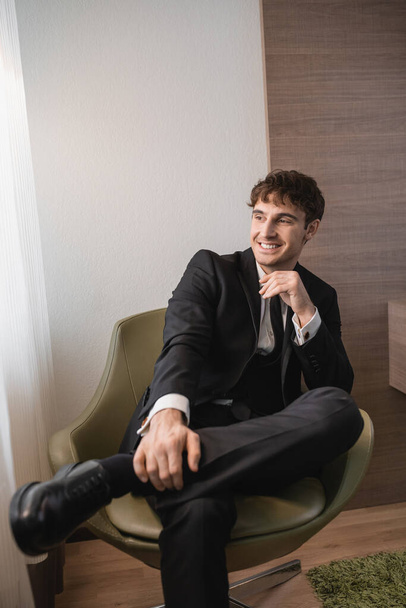 happy man in black formal wear with tie and classic shoes sitting on comfortable armchair and looking away on wedding day, resting in modern hotel room, good looking groom  - Photo, Image