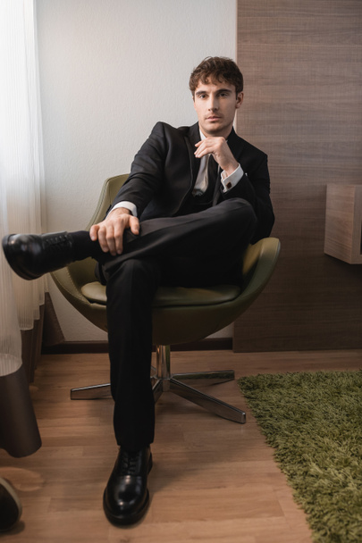 młody mężczyzna w czarnym stroju formalnym z krawatem i klasycznymi butami siedzący na wygodnym fotelu i patrzący w kamerę w dniu ślubu, pozujący w nowoczesnym pokoju hotelowym, przystojny pan młody  - Zdjęcie, obraz