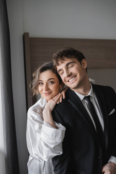 happy bride in jewelry and white silk robe leaning on shoulder of joyful groom in classic black suit while standing together in modern hotel room during honeymoon, newlyweds  - Fotó, kép