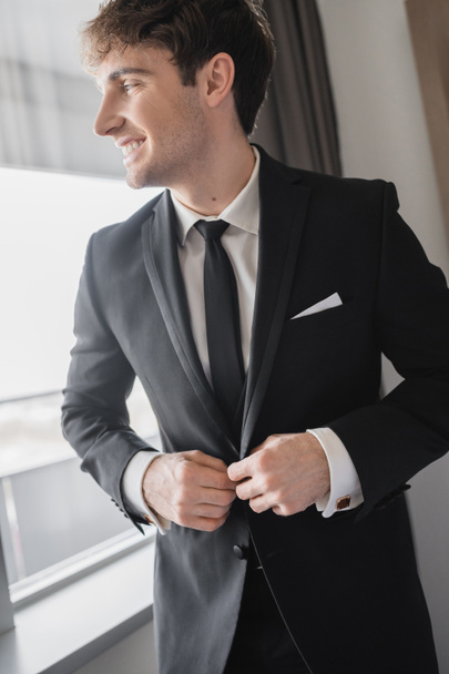 happy man in classic formal wear with black tie and white shirt buttoning blazer and standing in modern hotel room near window, groom on wedding day, special occasion   - Foto, Bild