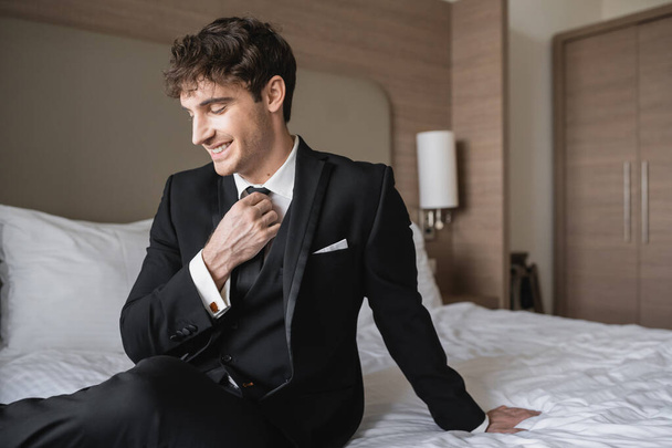 cheerful man in classy formal wear with white shirt adjusting black tie while looking away and sitting on bed in modern hotel room, groom on wedding day, special occasion   - Фото, изображение