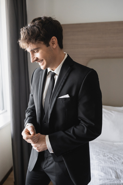 Porträt eines glücklichen jungen Mannes im klassischen schwarzen Anzug mit Krawatte und weißem Hemd, der im modernen Hotelzimmer lächelt, Bräutigam am Hochzeitstag, besonderer Anlass   - Foto, Bild