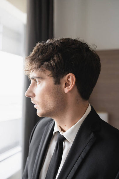 πορτρέτο του όμορφου άνδρα σε αριστοκρατικό επίσημο ένδυμα με μαύρη γραβάτα και λευκό πουκάμισο κοιτάζοντας το παράθυρο στο σύγχρονο δωμάτιο του ξενοδοχείου, γαμπρός την ημέρα του γάμου, ειδική περίσταση, πλαϊνή άποψη  - Φωτογραφία, εικόνα