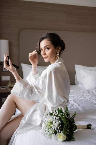 mujer atractiva en bata de seda blanca preparándose para su boda mientras hace maquillaje y sostiene el espejo de bolsillo, sentado en la cama cerca del ramo de novia en la habitación de hotel, ocasión especial, novia joven - Foto, Imagen