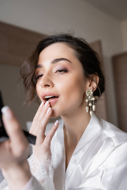 young bride with brunette hair in white silk robe preparing for her wedding while touching lips, holding pocket mirror in hotel room on wedding day, special occasion,  - Фото, зображення