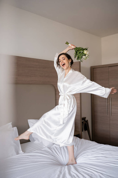 izgatott fiatal menyasszony barna haj fehér selyem köntös és gyöngy fülbevaló gazdaság menyasszonyi csokor, miközben ugrál az ágyon a szállodai szobában, különleges alkalom, boldog nő  - Fotó, kép