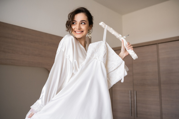 happy young bride with brunette hair in white silk robe holding soft hanger with elegant wedding dress and smiling in bedroom of hotel room, special occasion, charming woman  - Valokuva, kuva