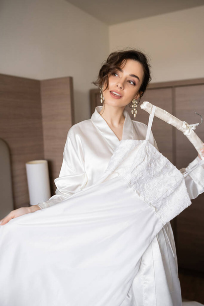 young bride with brunette hair in white silk robe holding soft hanger with elegant wedding dress in bedroom of hotel room, special occasion, charming woman  - Foto, Bild