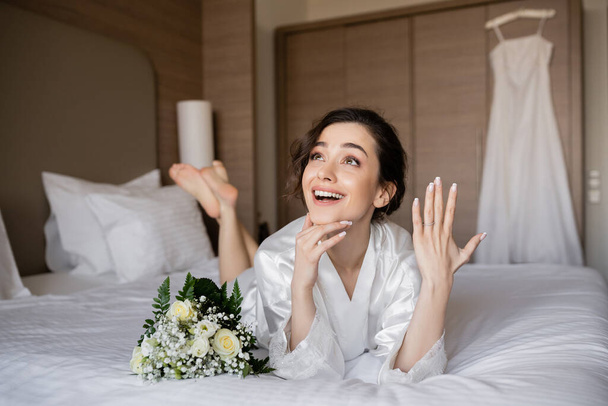 donna felice con capelli castani sdraiata vestaglia di seta bianca e mostrando l'anello di fidanzamento al dito accanto al bouquet da sposa sul letto in camera d'albergo con abito da sposa su sfondo sfocato, giovane sposa - Foto, immagini