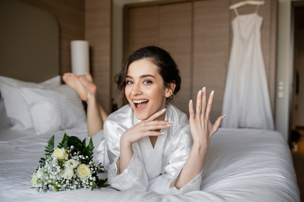 изумленная женщина с брюнетками в белом шелковом халате и с обручальным кольцом на пальце рядом с свадебным букетом на кровати в гостиничном номере с свадебным платьем на размытом фоне, молодая невеста - Фото, изображение
