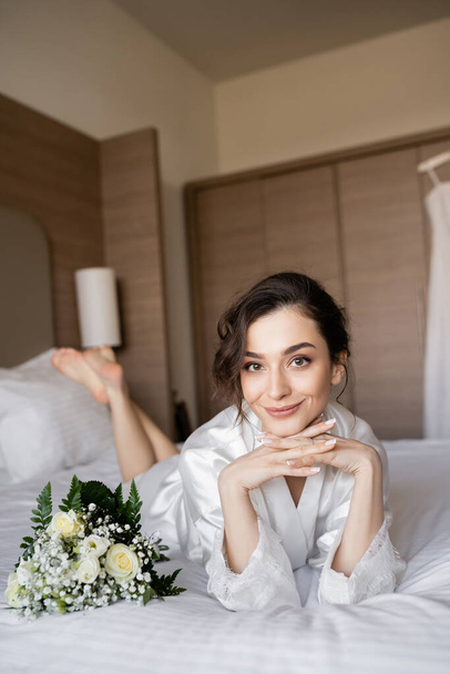 charmante vrouw met brunette haar liggend in witte zijden mantel met gebalde handen naast bruidsboeket op bed in hotelkamer op trouwdag, speciale gelegenheid, jonge bruid - Foto, afbeelding