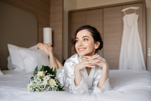 vrolijke vrouw met brunette haar liggend in witte zijden mantel met gebalde handen naast bruidsboeket op bed in hotelkamer met trouwjurk op wazige achtergrond, speciale gelegenheid, jonge bruid - Foto, afbeelding