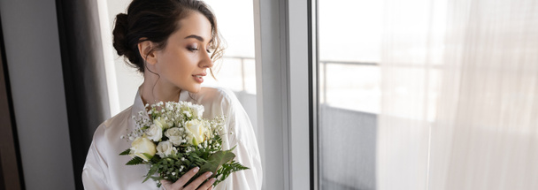 молодая женщина с брюнетками, стоящая в белом шелковом халате и держащая свадебный букет рядом с тюлем занавес и окно в номере отеля, особый случай, невеста в день свадьбы, баннер  - Фото, изображение