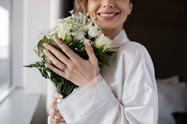 vista cortada de noiva feliz com anel de noivado no dedo de pé em roupão de seda branca e segurando buquê de noiva ao lado da janela na suíte do hotel, ocasião especial  - Foto, Imagem