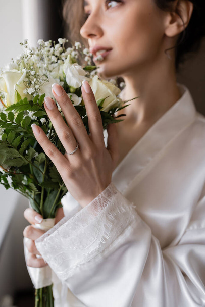 blurred bride with engagement ring on finger standing in white silk robe and holding bridal bouquet in modern hotel suite on wedding day, special occasion  - Photo, image